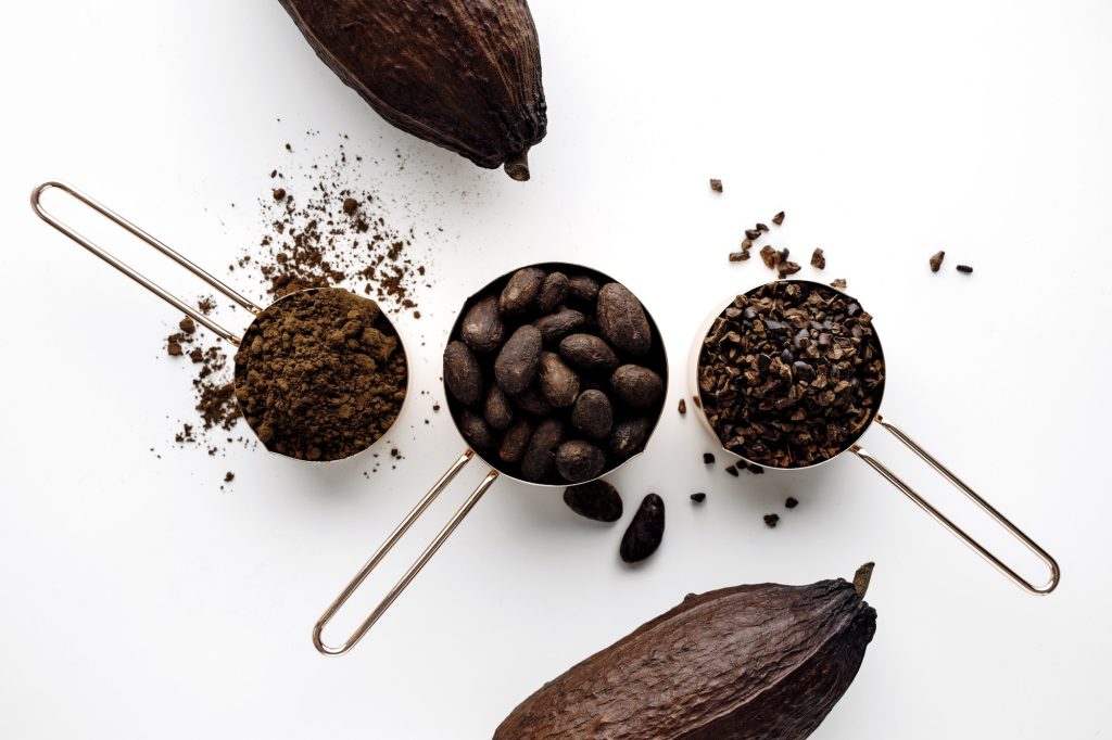 Flat lay rose gold measuring cups of cocoa beans, cacao nibs, cocoa powder and cocoa pods.